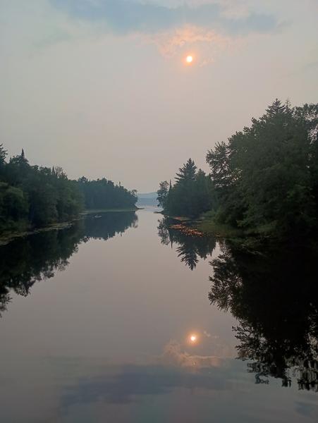 An eerie orange sun in a grey sky over the mirror-like Piseco Outlet on a hot day.